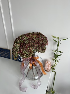 A bouquet of hydrangeas with faded green and pinkish-red petals is placed in a clear glass vase tied with orange and lavender ribbon. The vase sits on a white surface. A pink Himalayan salt lamp and a bamboo plant are nearby. The setting includes a motivational sign on the wall with a dark blue background and white text.