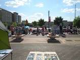 A bustling outdoor market scene with multiple stalls and people interacting under a clear blue sky. Vendors display various items, such as crafts and art, on tables. Shoppers browse the stalls, framed by urban buildings and trees in the background. A decorative flower arrangement is present in the foreground.