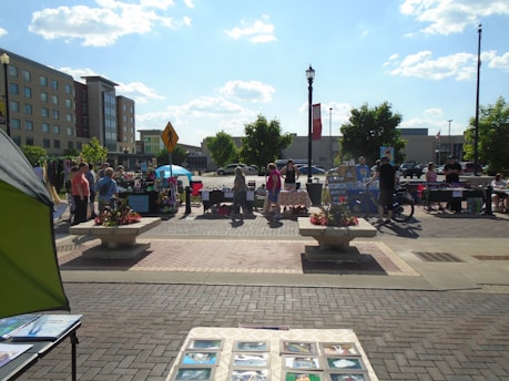 A bustling outdoor market scene with multiple stalls and people interacting under a clear blue sky. Vendors display various items, such as crafts and art, on tables. Shoppers browse the stalls, framed by urban buildings and trees in the background. A decorative flower arrangement is present in the foreground.