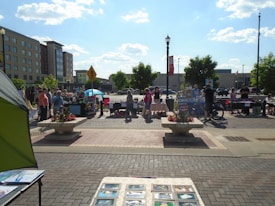 A bustling outdoor market scene with multiple stalls and people interacting under a clear blue sky. Vendors display various items, such as crafts and art, on tables. Shoppers browse the stalls, framed by urban buildings and trees in the background. A decorative flower arrangement is present in the foreground.