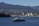 A modern ferry is cruising through a wide expanse of water, with a backdrop of industrial buildings and cranes. On the horizon, densely forested hills and distant mountains create a layered landscape. A large cargo ship can be seen near the shoreline, adding a juxtaposition of nature and industry to the scene.