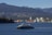 A modern ferry is cruising through a wide expanse of water, with a backdrop of industrial buildings and cranes. On the horizon, densely forested hills and distant mountains create a layered landscape. A large cargo ship can be seen near the shoreline, adding a juxtaposition of nature and industry to the scene.