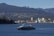 A modern ferry is cruising through a wide expanse of water, with a backdrop of industrial buildings and cranes. On the horizon, densely forested hills and distant mountains create a layered landscape. A large cargo ship can be seen near the shoreline, adding a juxtaposition of nature and industry to the scene.