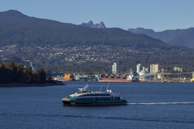 A modern ferry is cruising through a wide expanse of water, with a backdrop of industrial buildings and cranes. On the horizon, densely forested hills and distant mountains create a layered landscape. A large cargo ship can be seen near the shoreline, adding a juxtaposition of nature and industry to the scene.