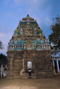 A historical temple structure with intricate carvings and sculptures decorates the facade. The tower features multiple levels adorned with figures and detailed artwork, painted in shades of blue, green, and stone. The entrance is situated centrally, and the surrounding area includes sparse trees and some ancient-looking walls.