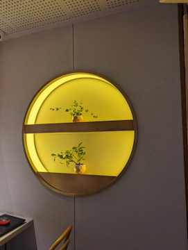 A circular wall installation features two shelves illuminated with a warm yellow light. Each shelf holds a small decorative plant in a pot. The installation is mounted on a gray textured wall, and part of an office setup with a black mouse on a desk is visible in the bottom left corner.
