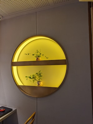 A circular wall installation features two shelves illuminated with a warm yellow light. Each shelf holds a small decorative plant in a pot. The installation is mounted on a gray textured wall, and part of an office setup with a black mouse on a desk is visible in the bottom left corner.