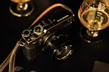 A vintage-style camera is placed on a dark surface, surrounded by two glasses containing a golden liquid. The camera has a leather strap and visible dials and buttons, suggesting a manual model. The setting implies a cozy, possibly dimly lit ambiance, emphasized by the warm, golden tones from the glass contents.