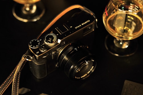A vintage-style camera is placed on a dark surface, surrounded by two glasses containing a golden liquid. The camera has a leather strap and visible dials and buttons, suggesting a manual model. The setting implies a cozy, possibly dimly lit ambiance, emphasized by the warm, golden tones from the glass contents.