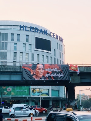 A cityscape featuring the Hledan Centre building with a large banner displaying a person's portrait and text in a foreign script. The scene includes a busy street with cars and motorbikes passing by. The sky is a light shade, indicating early morning or late afternoon.