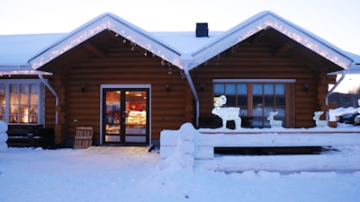 A cozy log cabin building is adorned with festive string lights along the roof. Outside, there is snow on the ground and a few illuminated reindeer decorations are placed on the porch. The cabin has large windows with warm lighting visible inside, suggesting a welcoming atmosphere. There is a wooden crate leaning against the wall near the entrance.