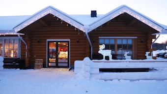 A cozy log cabin building is adorned with festive string lights along the roof. Outside, there is snow on the ground and a few illuminated reindeer decorations are placed on the porch. The cabin has large windows with warm lighting visible inside, suggesting a welcoming atmosphere. There is a wooden crate leaning against the wall near the entrance.