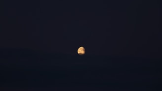 A bright, partially illuminated moon rises against a dark night sky. The moon's surface features are visible, with soft shadows and highlights creating a three-dimensional appearance. The rest of the scene is enveloped in darkness, emphasizing the glowing lunar surface.