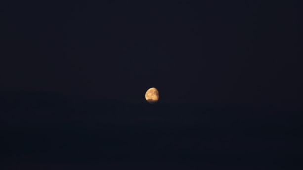 A bright, partially illuminated moon rises against a dark night sky. The moon's surface features are visible, with soft shadows and highlights creating a three-dimensional appearance. The rest of the scene is enveloped in darkness, emphasizing the glowing lunar surface.
