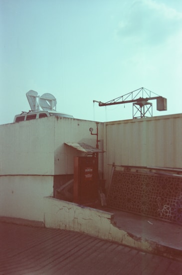 A rooftop area with a partially visible vehicle topped with satellite dishes, an industrial crane in the background, and a red utility box next to a patterned metal grill leaning against a wall.
