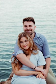A couple sits closely together on a rock by the water, with the man embracing the woman from behind. They both wear casual clothing, with the woman in a light blue outfit and the man in a darker blue shirt. Both are smiling gently, conveying a sense of warmth and affection.