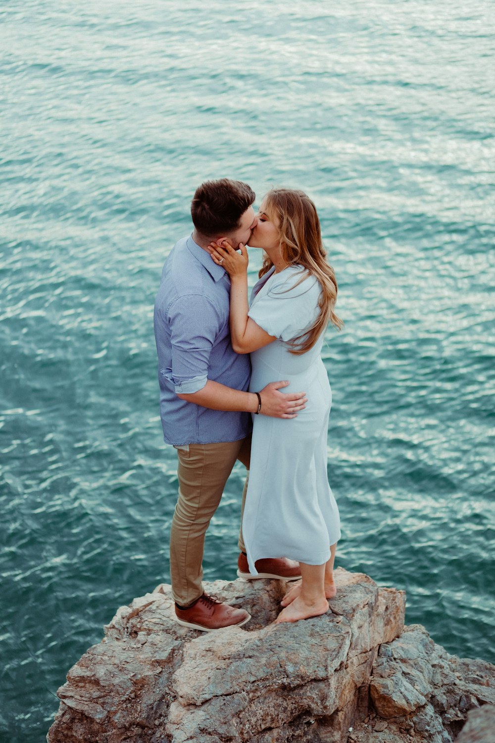 a man and woman kissing on a rock by the water
