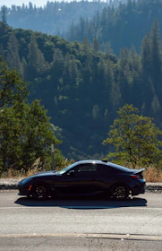 A sleek black luxury car parked beside a lush green mountain road at sunrise.