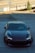 A sleek, dark-colored sports car is parked on a paved area near an overpass. The car's headlights are on, and a person is visible inside the vehicle, wearing a white shirt. In the background, there's a sign that includes directions towards the American River. The scenery also includes some greenery and a bridge overpass.