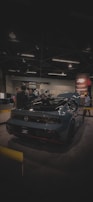 A sleek, modern sports car with a glossy dark finish is on display at an indoor exhibition. Several people are gathered around it, engaging in conversation. The venue is dimly lit, with overhead lights casting a soft glow. A promotional banner is visible in the background.