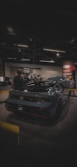 A sleek, modern sports car with a glossy dark finish is on display at an indoor exhibition. Several people are gathered around it, engaging in conversation. The venue is dimly lit, with overhead lights casting a soft glow. A promotional banner is visible in the background.