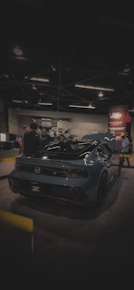 A sleek, modern sports car with a glossy dark finish is on display at an indoor exhibition. Several people are gathered around it, engaging in conversation. The venue is dimly lit, with overhead lights casting a soft glow. A promotional banner is visible in the background.