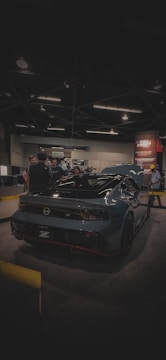 A sleek, modern sports car with a glossy dark finish is on display at an indoor exhibition. Several people are gathered around it, engaging in conversation. The venue is dimly lit, with overhead lights casting a soft glow. A promotional banner is visible in the background.