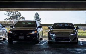 Two cars are parked side by side in a dimly lit parking garage. The car on the left is black, while the car on the right is white with distinctive headlights. Outside, trees and power lines are visible under a clear sky.
