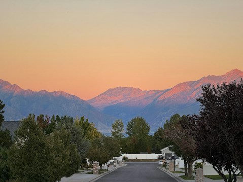 A neighborhood street lined with trees leads to a view of mountain ranges under a sky transitioning from soft orange to pale pink. The area appears quiet and residential, with well-kept lawns and clean sidewalks.