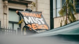 A person holds a large banner with bold text that reads 'TRUDEAU MUST RESIGN' in orange and white letters. The person appears to be in an outdoor urban setting with a building visible in the background. The scene includes some greenery including a tree and fence.