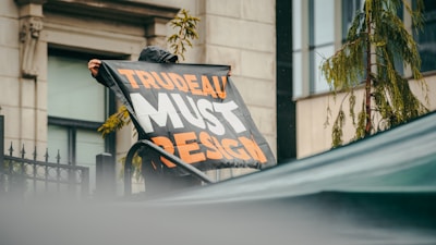 A person holds a large banner with bold text that reads 'TRUDEAU MUST RESIGN' in orange and white letters. The person appears to be in an outdoor urban setting with a building visible in the background. The scene includes some greenery including a tree and fence.