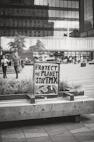 A black and white photograph features a sign with handwritten text that reads 'Protect the Planet Stop TMX' placed on a wooden bench surrounded by plants. The background shows a busy urban environment with modern office buildings and people walking.