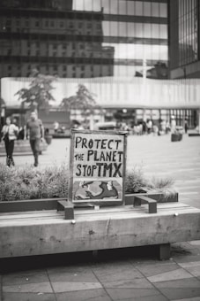 A black and white photograph features a sign with handwritten text that reads 'Protect the Planet Stop TMX' placed on a wooden bench surrounded by plants. The background shows a busy urban environment with modern office buildings and people walking.
