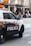 A police SUV with the words 'TRANSIT POLICE' boldly displayed on the side is parked on a city street. The vehicle is white and has emergency lights on top. In the background, blurred pedestrians are seen walking along the sidewalk, while store signs and street infrastructure are visible, suggesting a busy urban environment.