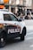 A police SUV with the words 'TRANSIT POLICE' boldly displayed on the side is parked on a city street. The vehicle is white and has emergency lights on top. In the background, blurred pedestrians are seen walking along the sidewalk, while store signs and street infrastructure are visible, suggesting a busy urban environment.