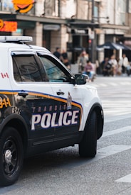 A police SUV with the words 'TRANSIT POLICE' boldly displayed on the side is parked on a city street. The vehicle is white and has emergency lights on top. In the background, blurred pedestrians are seen walking along the sidewalk, while store signs and street infrastructure are visible, suggesting a busy urban environment.