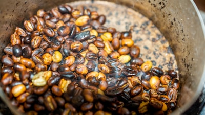 A close-up view of roasted coffee beans in a metal container. The beans are a mix of various shades of brown, indicating different roasting levels. The container has a scratched and worn appearance, with a gradient in color intensity from center to edge. There is a slight amount of smoke or steam visible, suggesting that the beans are freshly roasted.