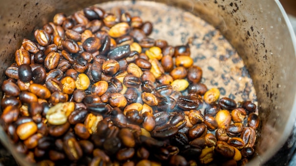 A close-up view of roasted coffee beans in a metal container. The beans are a mix of various shades of brown, indicating different roasting levels. The container has a scratched and worn appearance, with a gradient in color intensity from center to edge. There is a slight amount of smoke or steam visible, suggesting that the beans are freshly roasted.