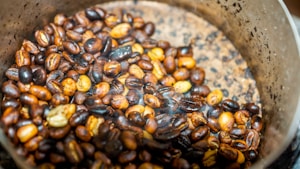 A close-up view of roasted coffee beans in a metal container. The beans are a mix of various shades of brown, indicating different roasting levels. The container has a scratched and worn appearance, with a gradient in color intensity from center to edge. There is a slight amount of smoke or steam visible, suggesting that the beans are freshly roasted.