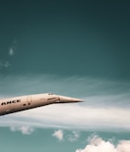 A sleek, supersonic airplane with a pointed nose is flying against a backdrop of a blue sky with scattered white clouds. The airplane has a metallic finish and the word 'ANCE' is partially visible on its side.