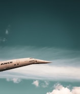 A sleek, supersonic airplane with a pointed nose is flying against a backdrop of a blue sky with scattered white clouds. The airplane has a metallic finish and the word 'ANCE' is partially visible on its side.