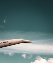 A sleek, supersonic airplane with a pointed nose is flying against a backdrop of a blue sky with scattered white clouds. The airplane has a metallic finish and the word 'ANCE' is partially visible on its side.