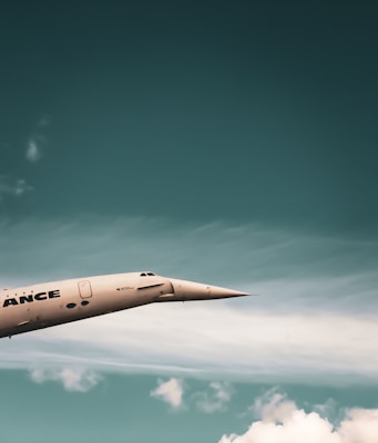 A sleek, supersonic airplane with a pointed nose is flying against a backdrop of a blue sky with scattered white clouds. The airplane has a metallic finish and the word 'ANCE' is partially visible on its side.