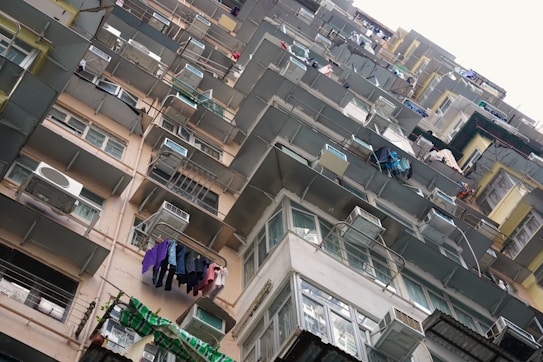 A high-rise residential building with numerous balconies and air conditioning units. Clothes are visible hanging to dry on several of the balconies. The building features a mix of beige, gray, and white tones, with some green and blue accents from the clothing.