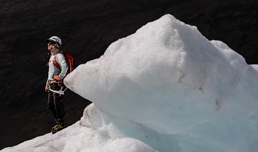 A person wearing a helmet and outdoor gear, including a red backpack, stands on a glacier. The individual appears to be prepared for ice climbing, with crampons on their boots. The glacier has a jagged, icy surface with a pale blue hue. In the background, dark mountain slopes can be seen.