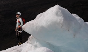 A person wearing a helmet and outdoor gear, including a red backpack, stands on a glacier. The individual appears to be prepared for ice climbing, with crampons on their boots. The glacier has a jagged, icy surface with a pale blue hue. In the background, dark mountain slopes can be seen.