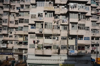 A densely-packed residential building with multiple stories featuring numerous windows and laundry hanging outside. The facade shows signs of wear and aging with faded paint and visible cracks.