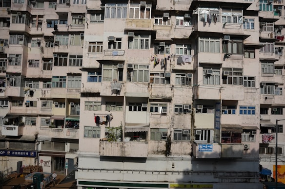 A densely-packed residential building with multiple stories featuring numerous windows and laundry hanging outside. The facade shows signs of wear and aging with faded paint and visible cracks.