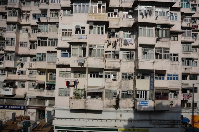 A densely-packed residential building with multiple stories featuring numerous windows and laundry hanging outside. The facade shows signs of wear and aging with faded paint and visible cracks.