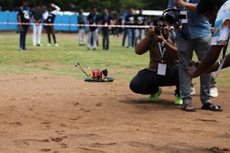 A drone flying close to the ground on a grassy field, surrounded by people. The drone has a red body and is being photographed by multiple photographers. Some people in the background are observing. The scene suggests a drone demonstration or competition gathering.